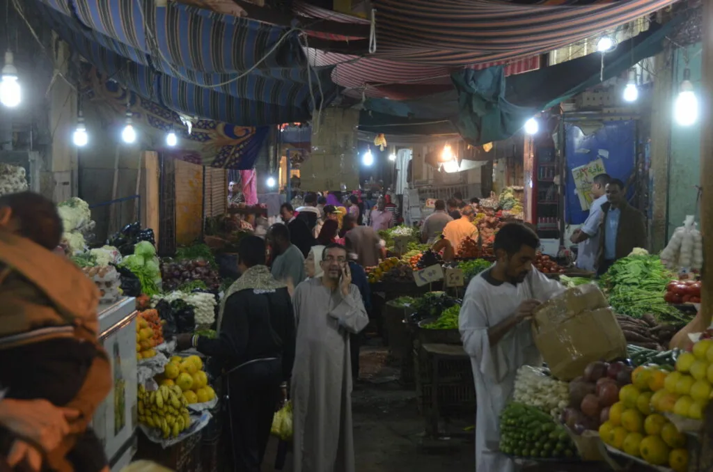 Crowded indoor market at night with produce stalls displaying colorful fruits and vegetables under striped awnings and bright lights