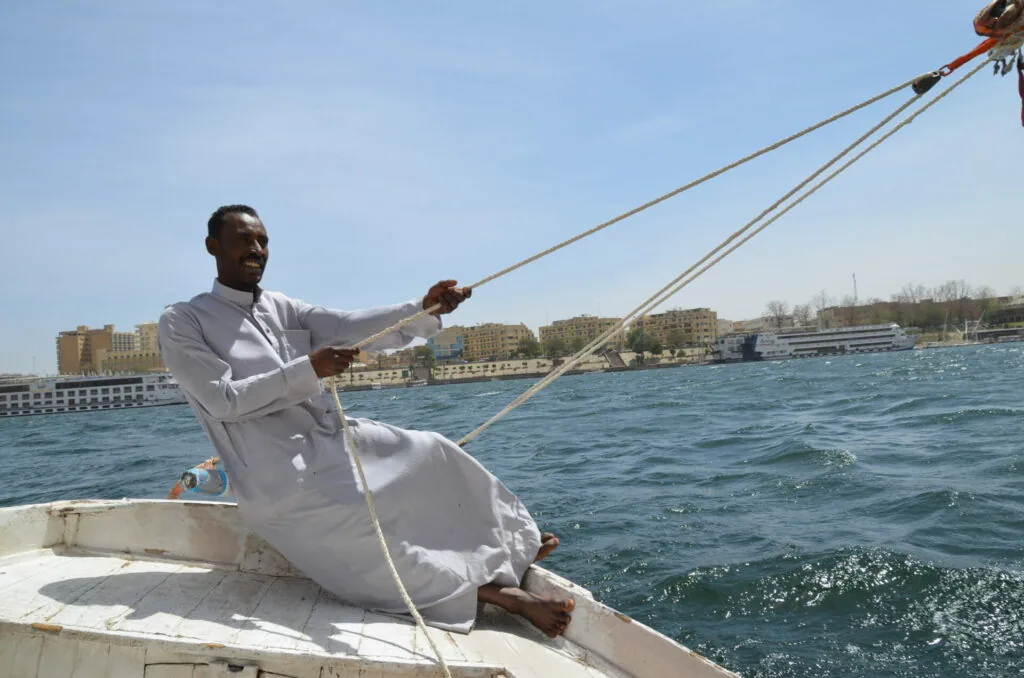 Man in white clothing holding boat rigging rope on a vessel in blue water with waterfront buildings in background