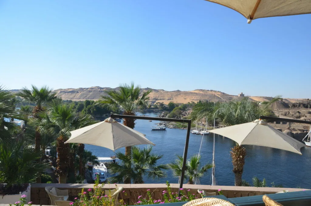 Waterfront terrace with canvas umbrellas, palm trees, and view of a river with boats and desert hills