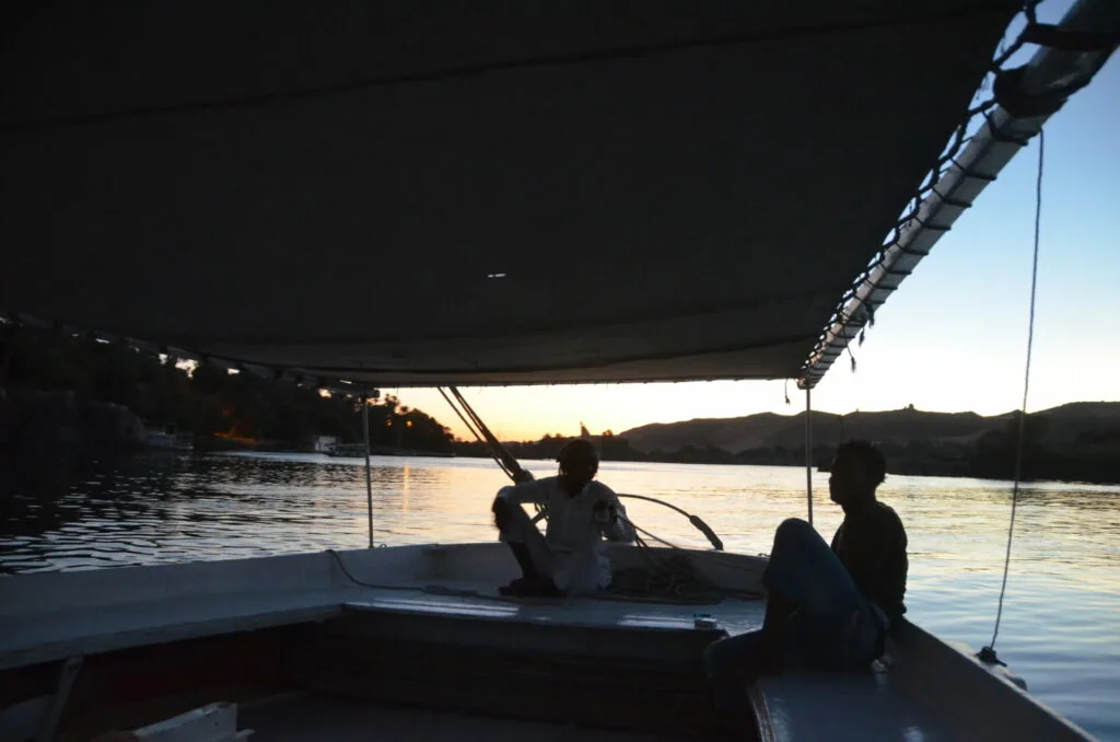 Silhouetted passengers sitting on a boat with a black canopy at sunset over calm water and distant hills