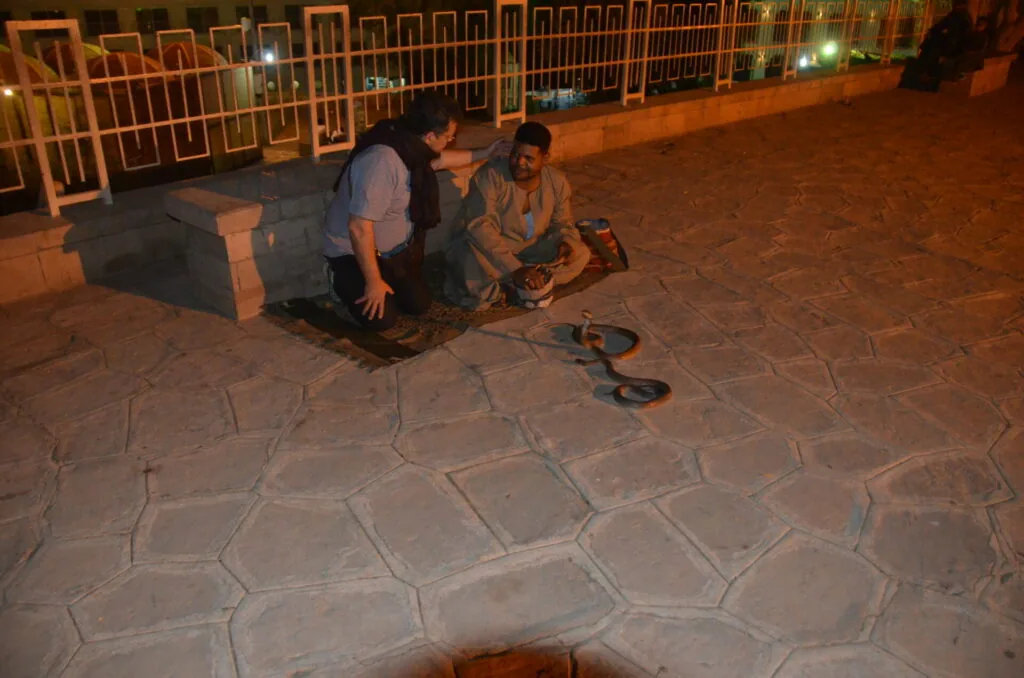 Two people crouching on paved ground at night examining skateboard equipment, with orange metal fencing visible in background