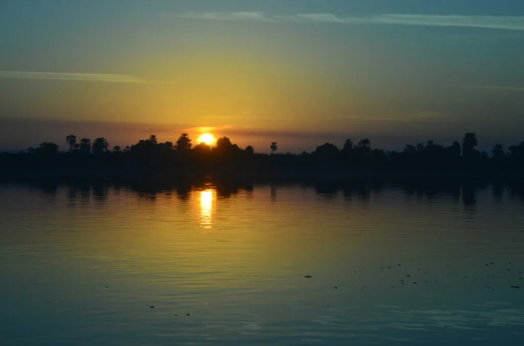 Sun rising over calm water with silhouetted trees along the shoreline at dawn.