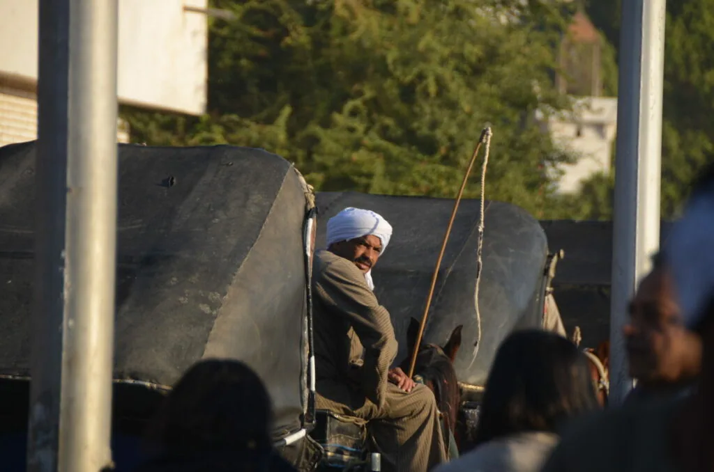 Person wearing white headwrap and tan clothing standing near large dark fabric or canvas with wooden poles, trees visible in background