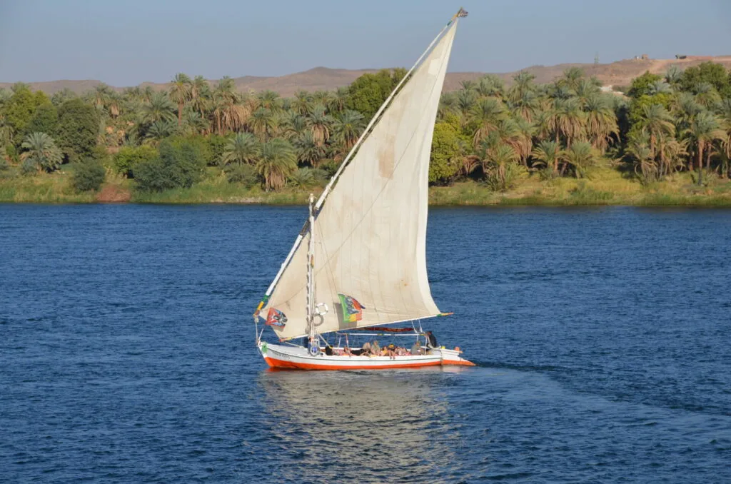 Traditional sailboat with white canvas sailing on blue water past palm-lined riverbank and desert hills