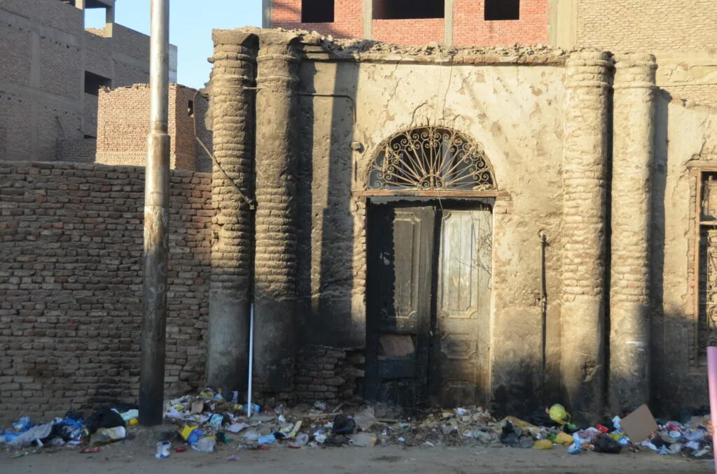 Deteriorated brick building with boarded entrance, debris scattered at base, surrounded by concrete walls
