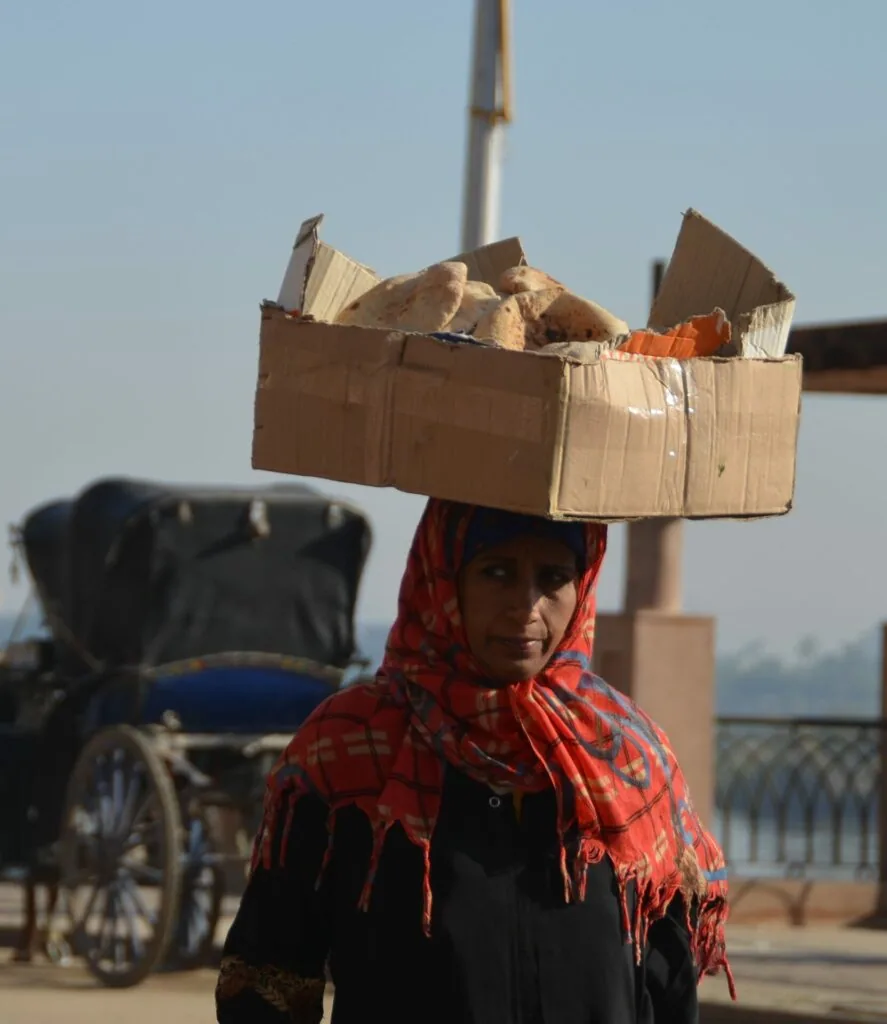 Person carrying large cardboard box filled with bread on head while wearing red patterned headscarf