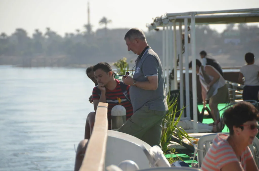 People gathered on a boat deck overlooking a river with palm trees and structures visible in the distant background.