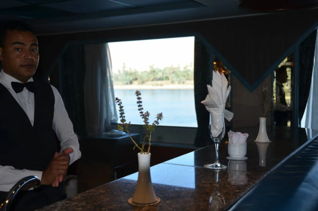 Man in formal attire standing behind a bar counter on a boat with water view through window behind him
