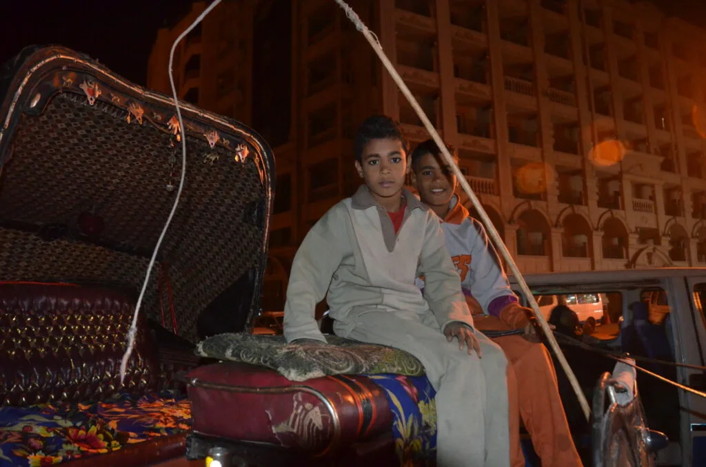 Two boys sitting in a decorated cart at night with buildings lit in warm tones behind them