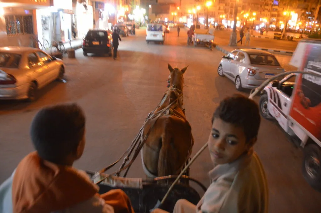 Two people riding in a horse-drawn cart on a busy city street at night with cars and pedestrians