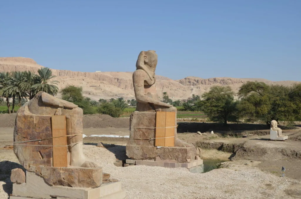 Ancient Egyptian stone statues and ruins in desert landscape with palm trees and river in background