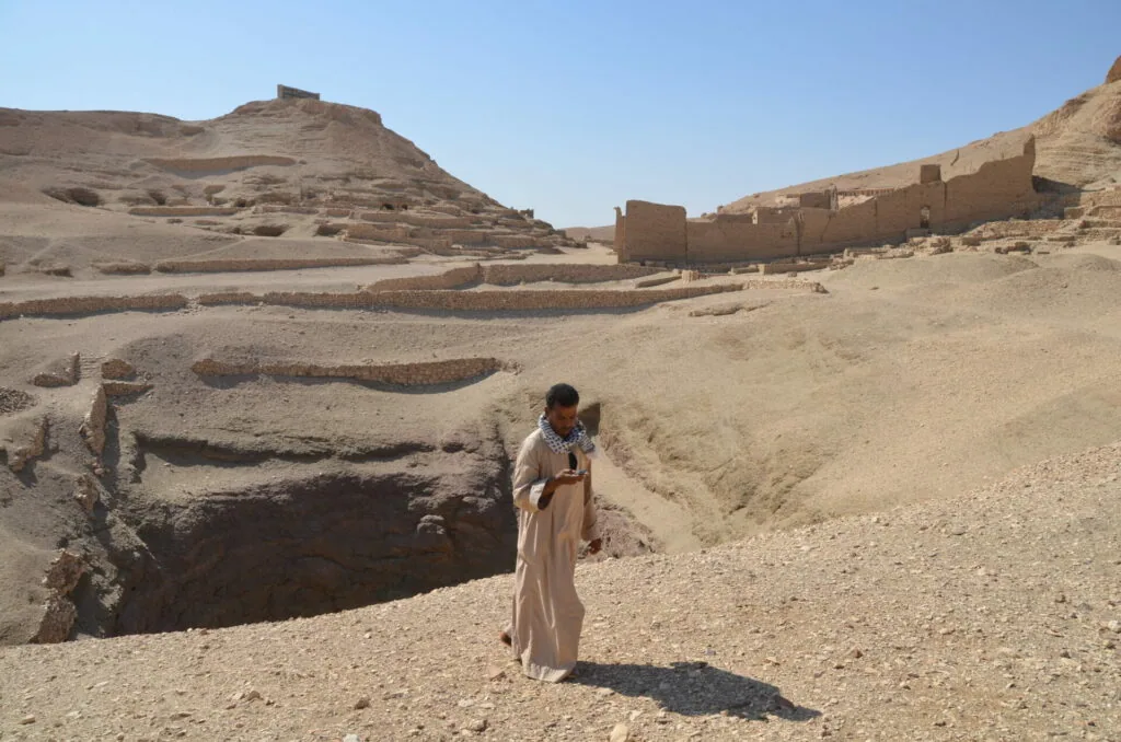 Person in light-colored clothing standing in desert archaeological site with excavated mud brick structures and hillside ruins