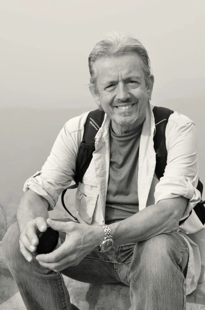 Smiling older man with gray hair wearing a light colored shirt and black vest, crouching against a neutral background