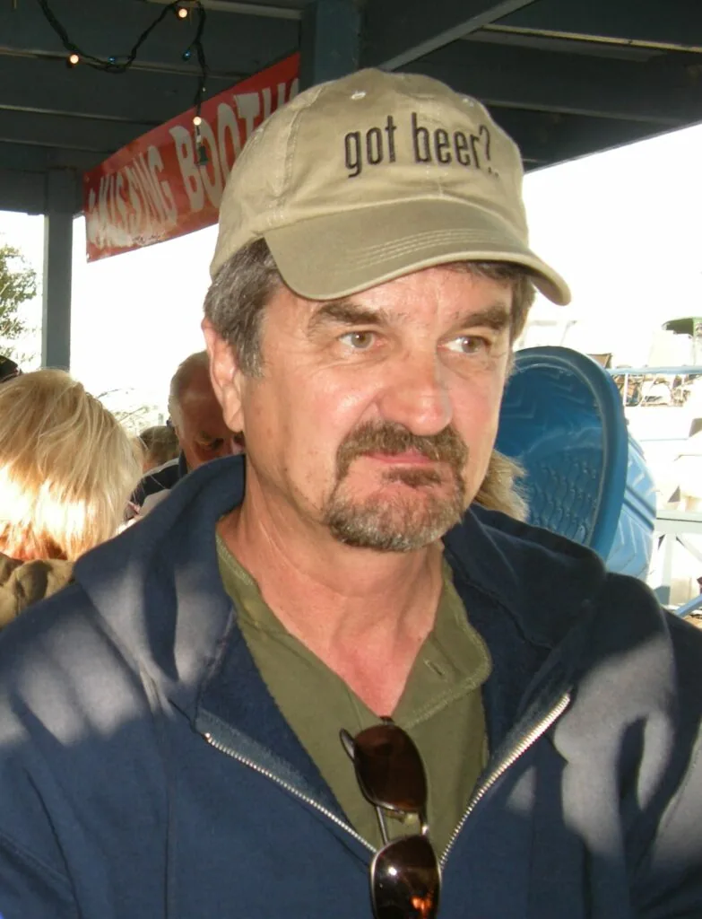 Man wearing tan baseball cap with "got beer?" text and blue jacket at outdoor venue