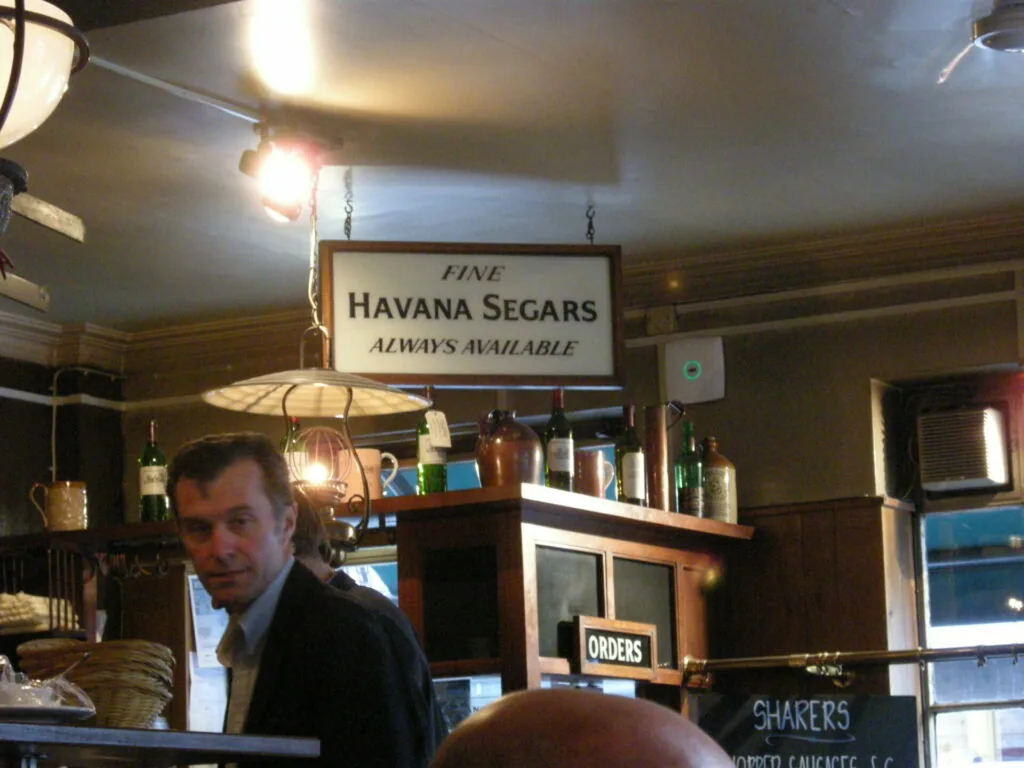 Man in dark jacket inside vintage bar with wooden paneling and hanging sign advertising fine Havana cigars