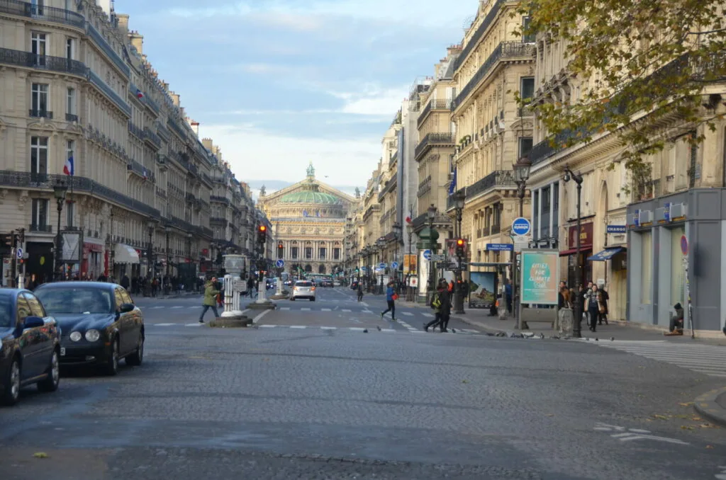 Parisian street with classical Haussmann buildings, pedestrians, and vehicles leading toward an ornate building with a green dome in the distance