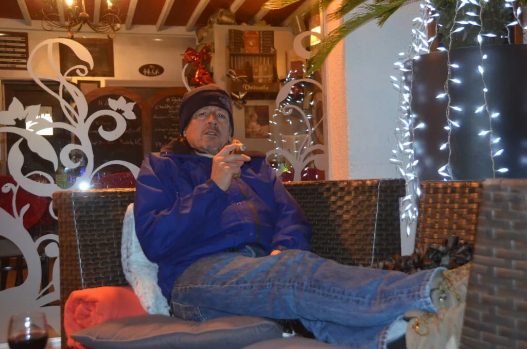 Man in blue jacket and cap sitting in wicker chair inside decorated venue with holiday lights and white floral wall art