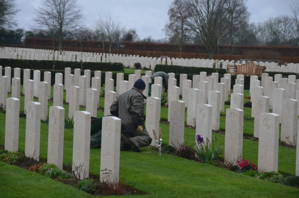 Person kneeling at white military headstone in cemetery surrounded by rows of gravestones and bare trees