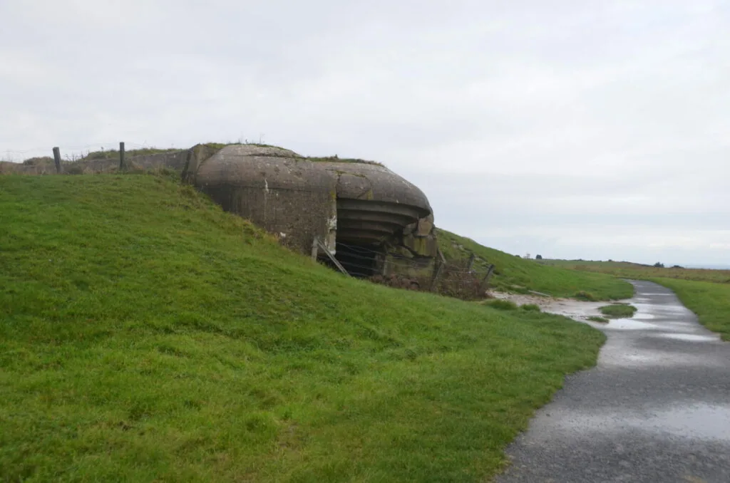 Abandoned concrete bunker embedded in grassy hillside with overgrown vegetation and narrow path leading to entrance