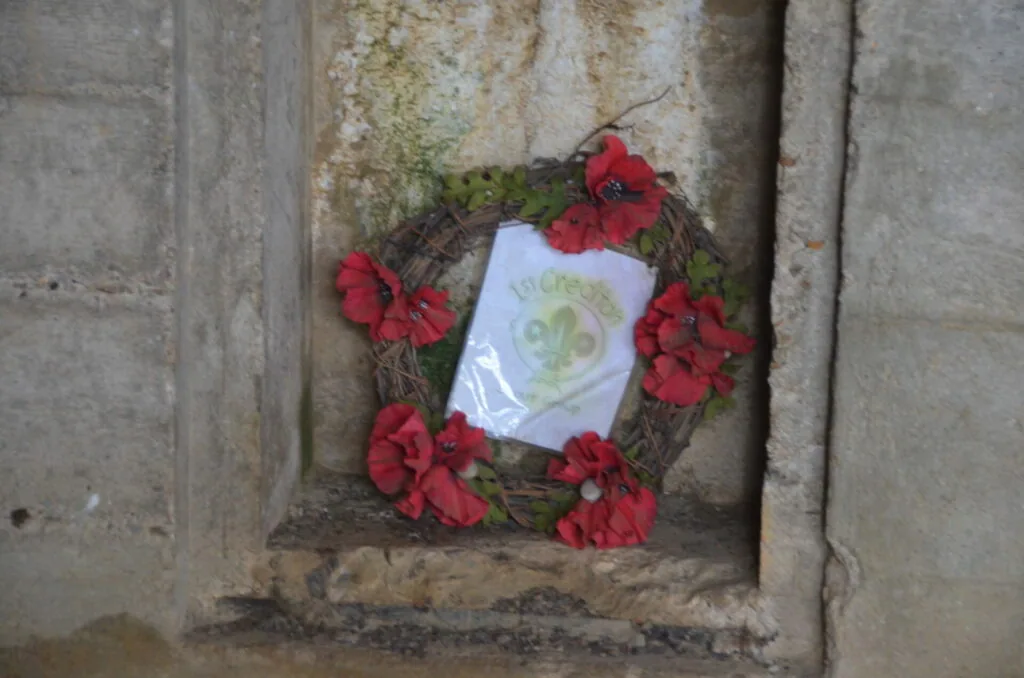 Wreath with red flowers and green foliage surrounding a white memorial plaque mounted on weathered stone wall