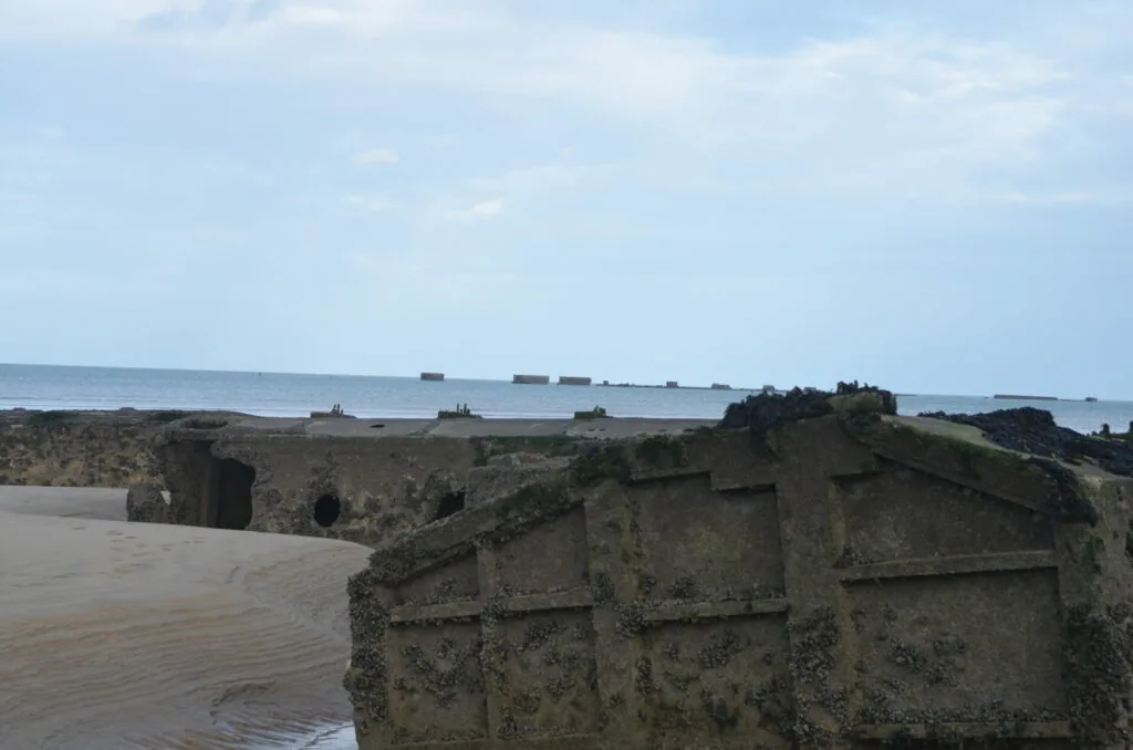 Concrete bunker ruins on sandy beach with ships visible on horizon under cloudy sky
