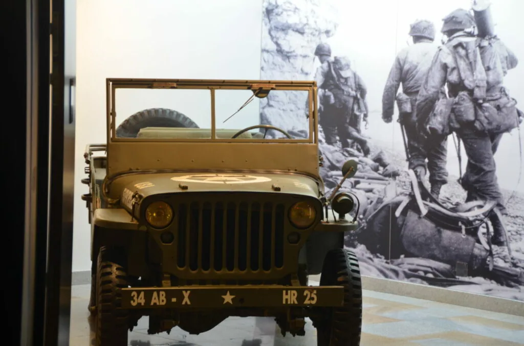 Vintage military Jeep with registration plate 34 AB-X HR 25 displayed in museum with black and white photograph of soldiers on wall behind it