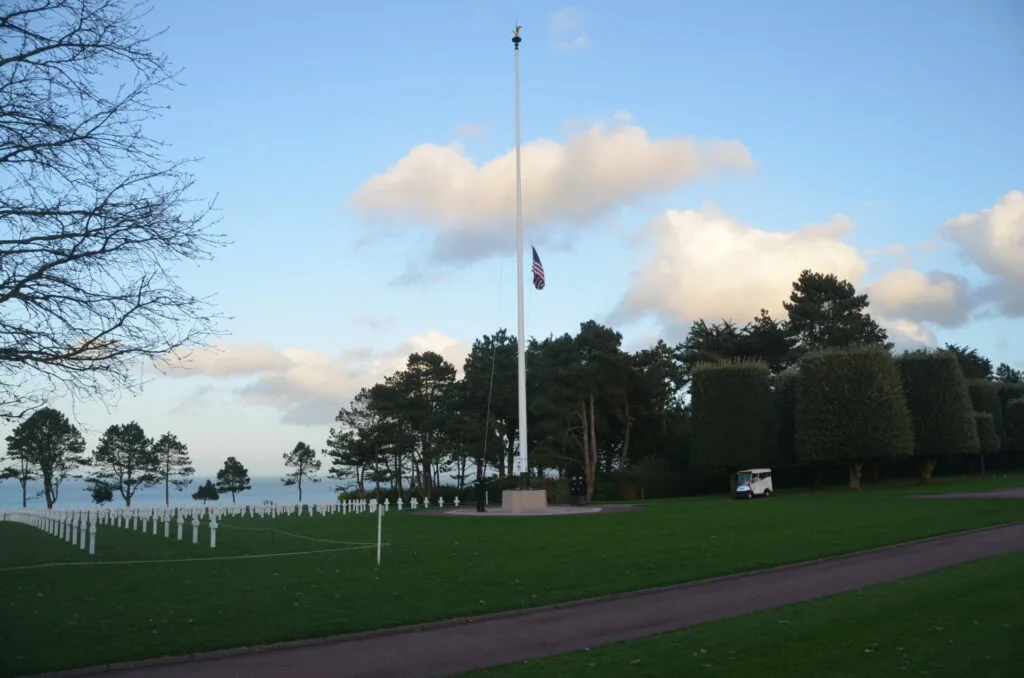 Military cemetery with white crosses, tall flagpole at half-mast, and evergreen trees under blue sky with clouds
