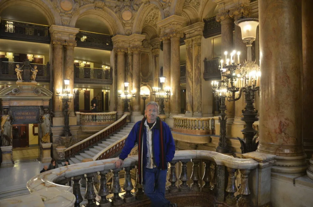 Man in blue jacket standing at ornate marble staircase in grand neoclassical building interior with columns, arches, and brass chandeliers.