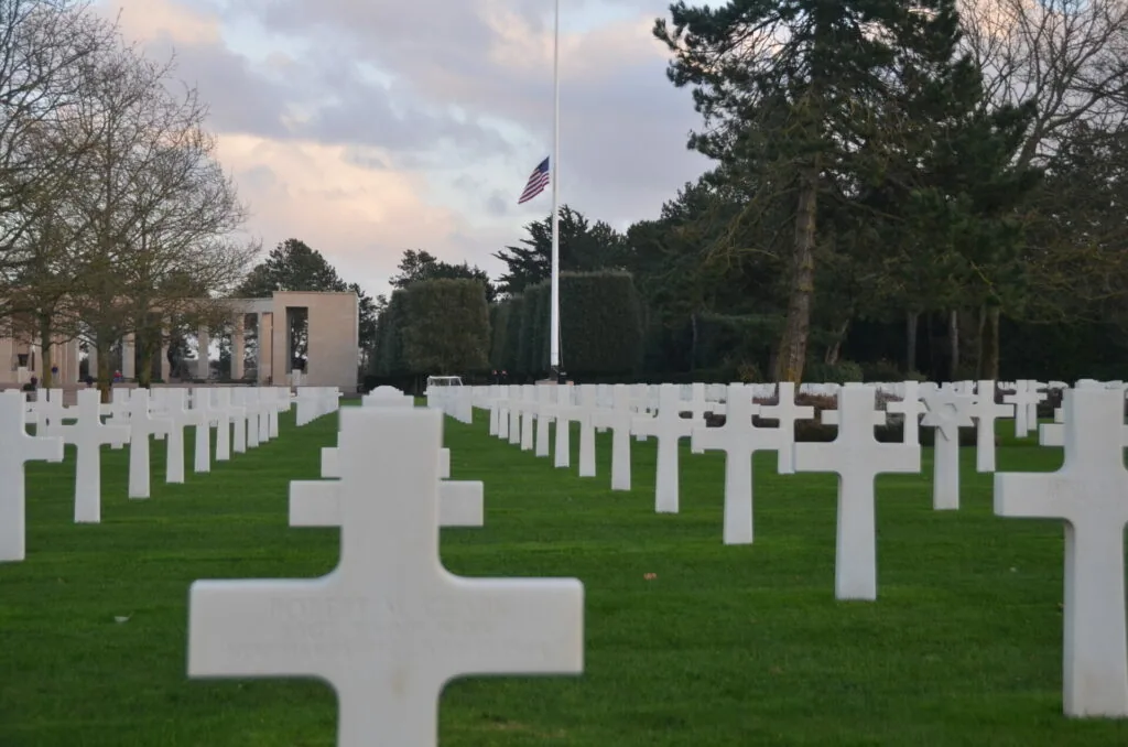 White military cemetery crosses arranged in rows on green grass with American flag and chapel building in background