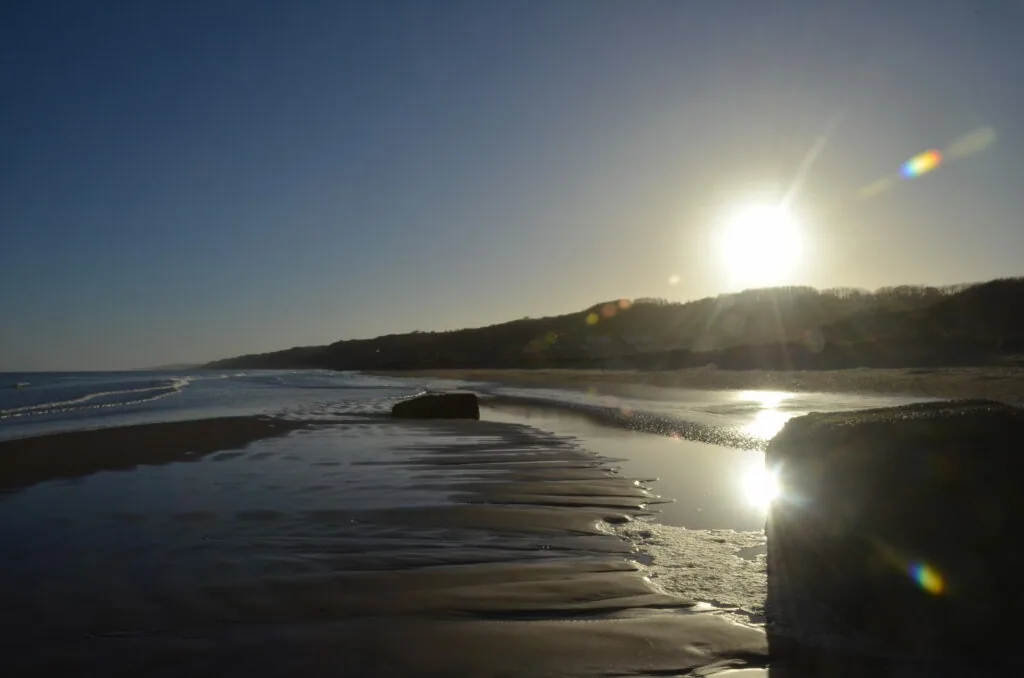 Coastal beach scene at sunset with rocky outcrops, calm waters, and forested hillside under bright sun