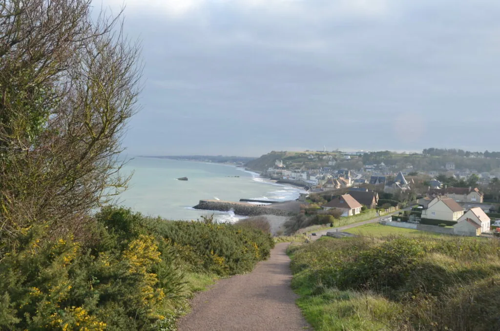Coastal village with church spire along rocky shoreline, viewed from hilltop path with flowering shrubs