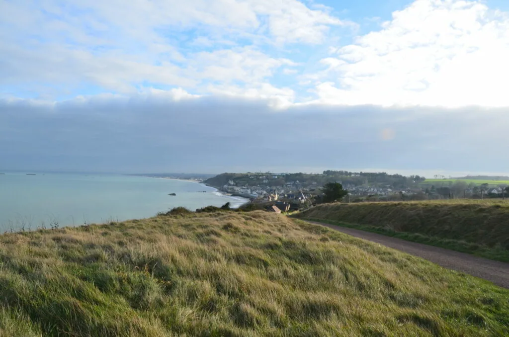 Coastal landscape with grassy hilltop overlooking a bay, seaside town, and agricultural fields under partly cloudy sky