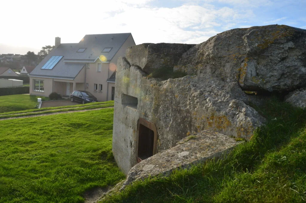 Concrete bunker with metal door in grassy field, modern house with solar panels visible in background