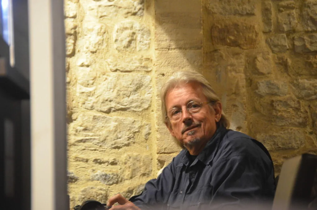Older man with light hair and goatee sitting against textured stone wall wearing dark shirt