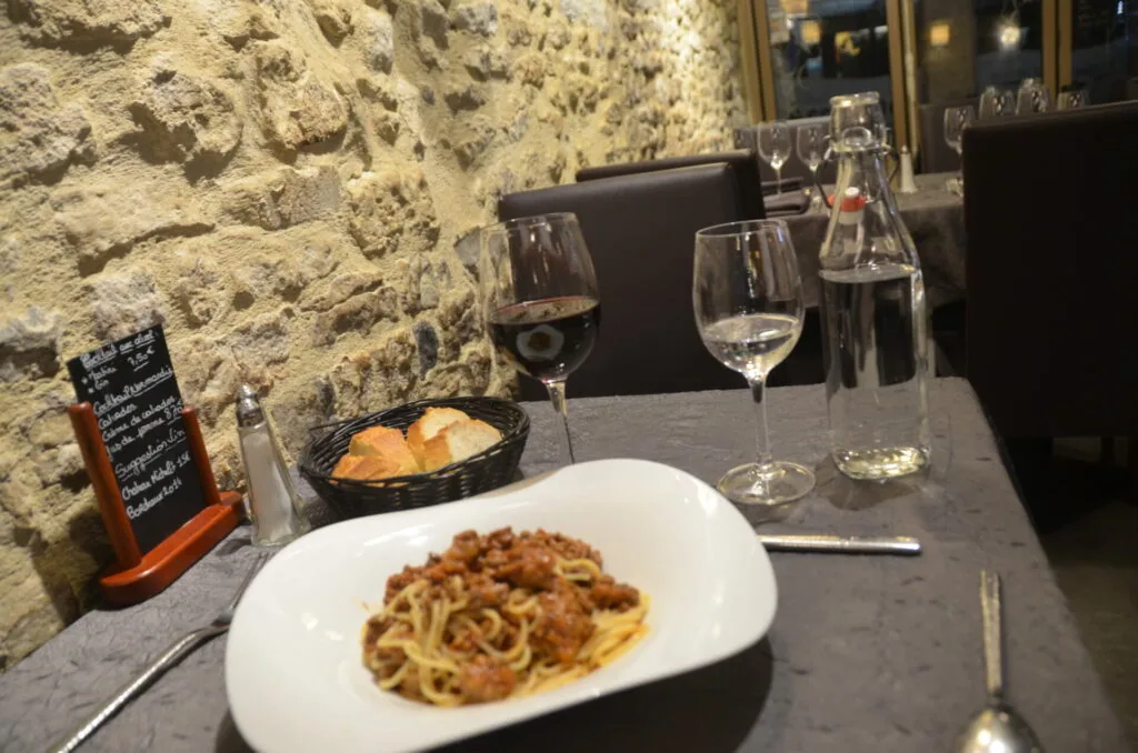 Pasta dish on white plate with wine glasses and bread basket on stone wall restaurant table