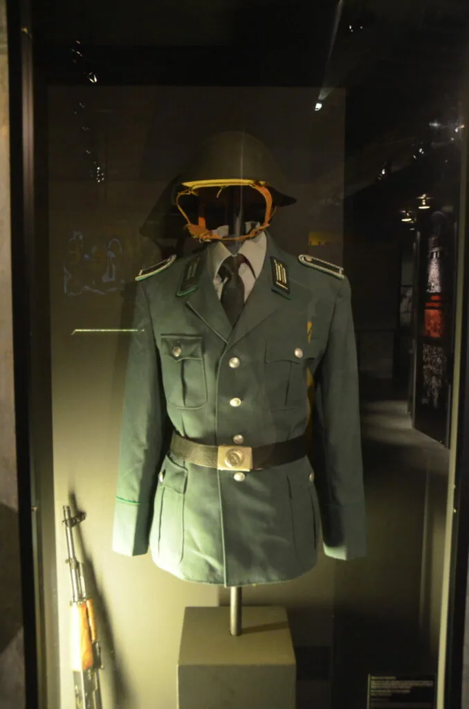 Military uniform display with green jacket, belt, tie, and woven cap on mannequin in museum case
