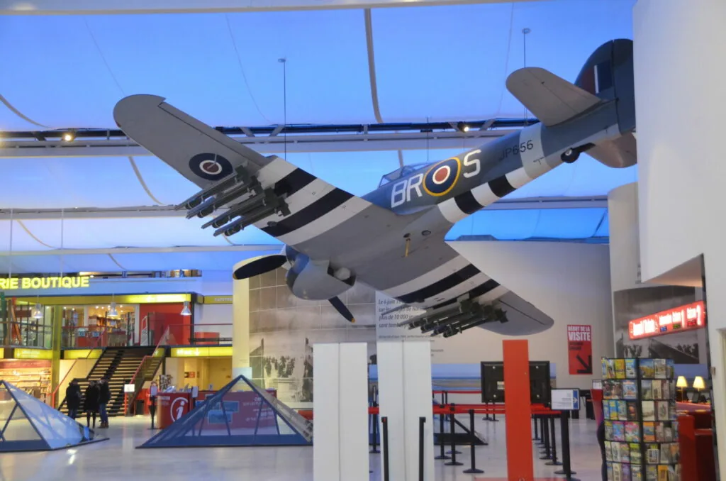 Military transport aircraft suspended from ceiling in museum exhibition space with retail storefronts below