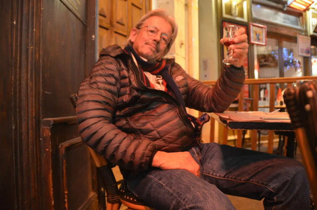 Man in brown puffer jacket and glasses raising a wine glass while seated indoors