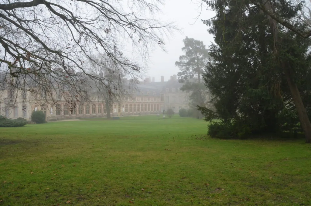 Grand estate with stone façade visible through mist across expansive lawn, bare tree branches in foreground and evergreen tree on right