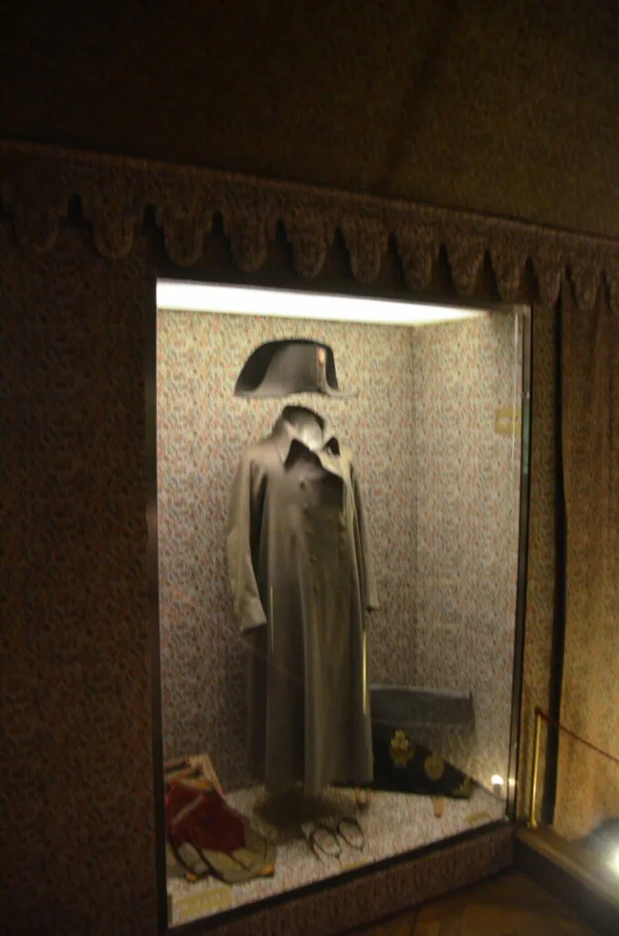Museum display case with illuminated mannequin wearing long coat and bicorne hat against patterned wallpaper background