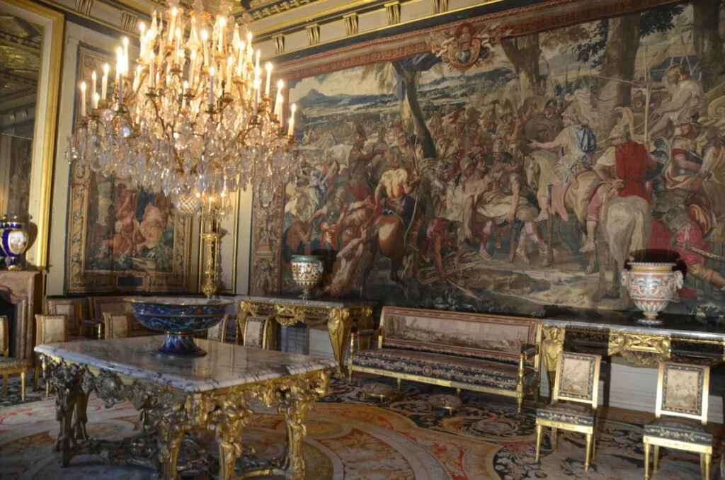Ornate palace room with crystal chandelier, gilded furniture, tapestries depicting classical scenes, and marble-topped tables