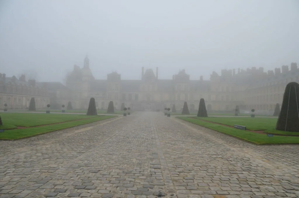 Formal courtyard with stone pathway, manicured lawns, topiary trees, and palatial architecture shrouded in fog