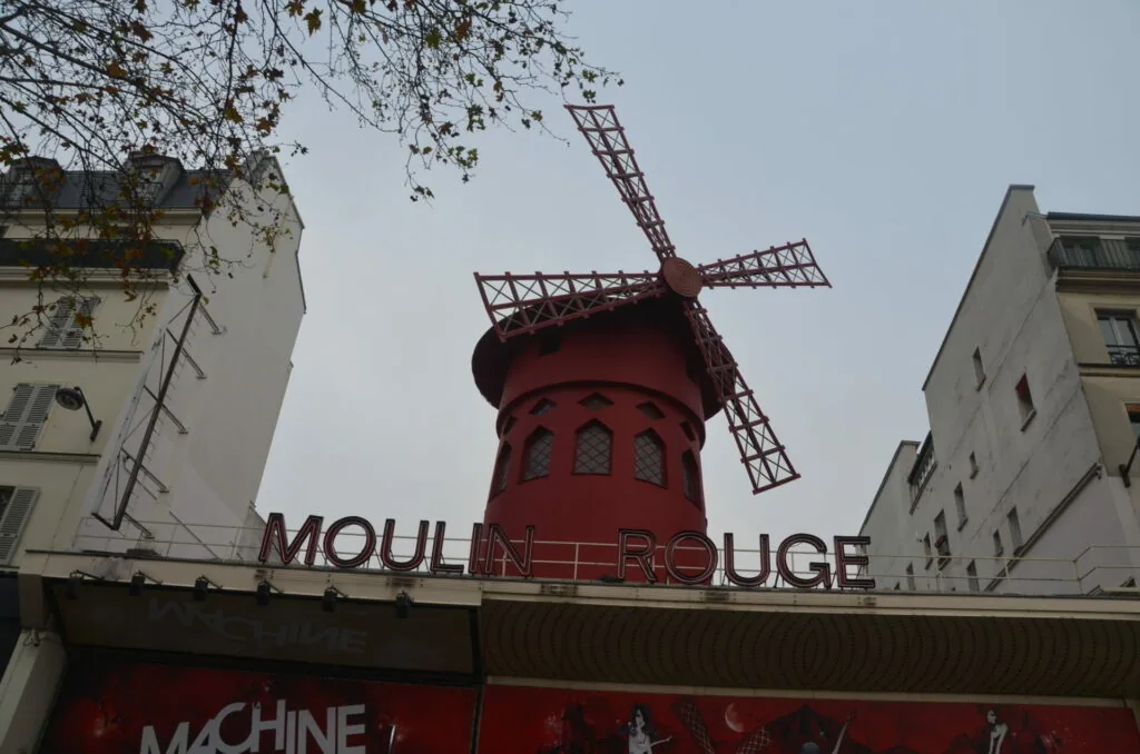 Red windmill with latticed sails perched atop a red building between Parisian apartment buildings