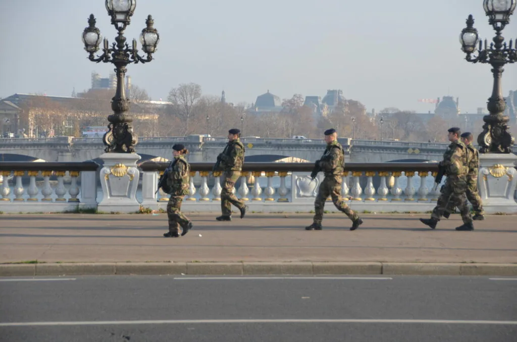 Military personnel in camouflage uniforms walking across a bridge with ornamental lampposts and classical balustrade, river and cityscape in background