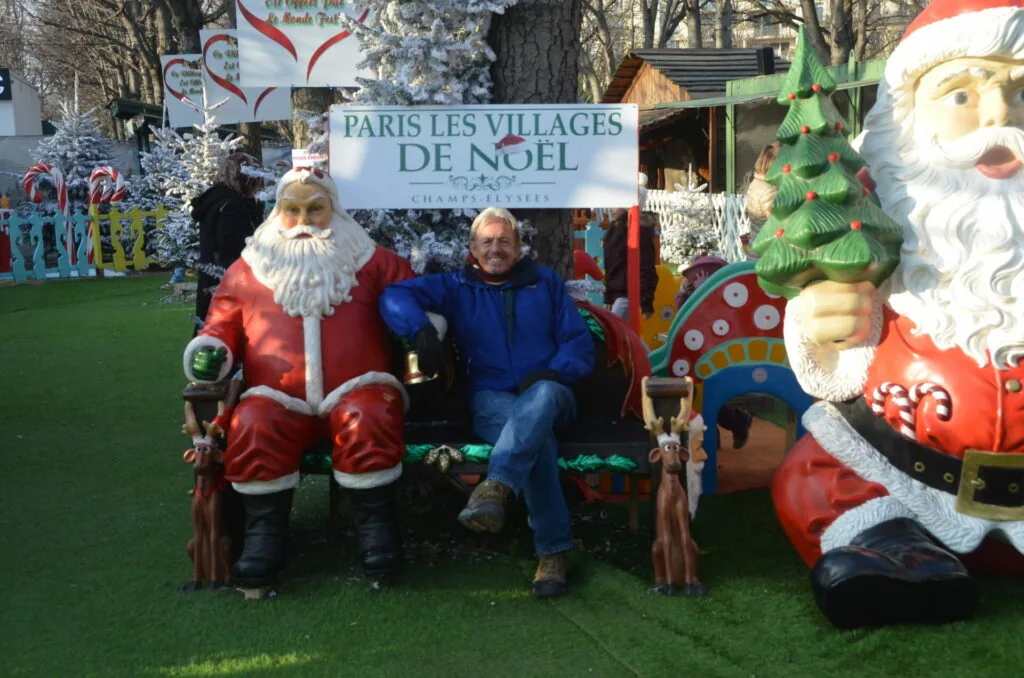 Man in blue jacket seated between two large Santa Claus sculptures at outdoor Christmas market