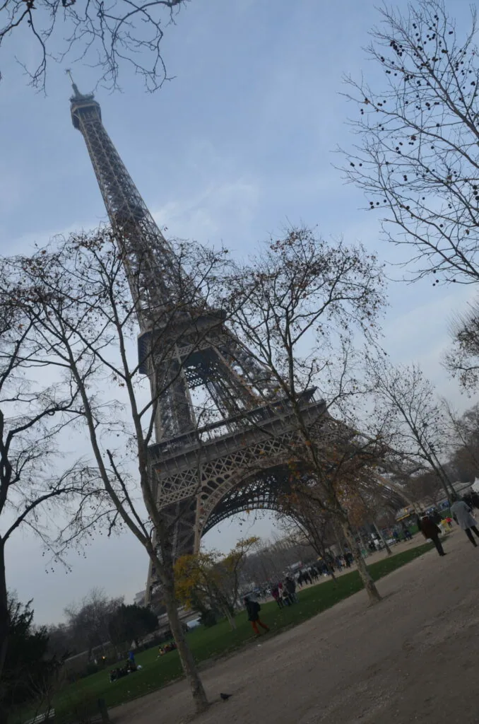 Iron lattice tower with ornate architecture framed by bare trees in a park setting with visitors below