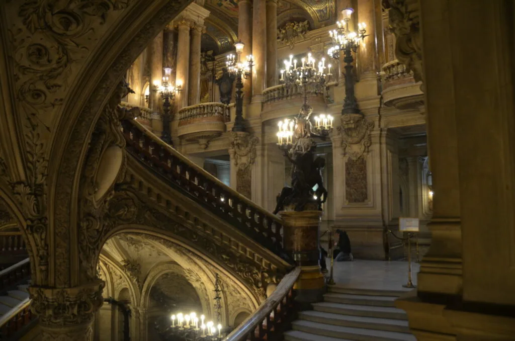 Grand ornate staircase with cream-colored walls, decorative molding, illuminated wall sconces, and crystal chandeliers in an elegant classical interior