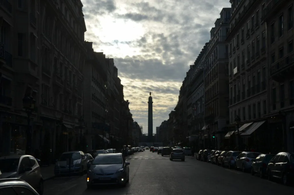 Parisian avenue with symmetrical Haussmann buildings, parked cars, and a tall column monument centered in the distance under dramatic cloudy sky