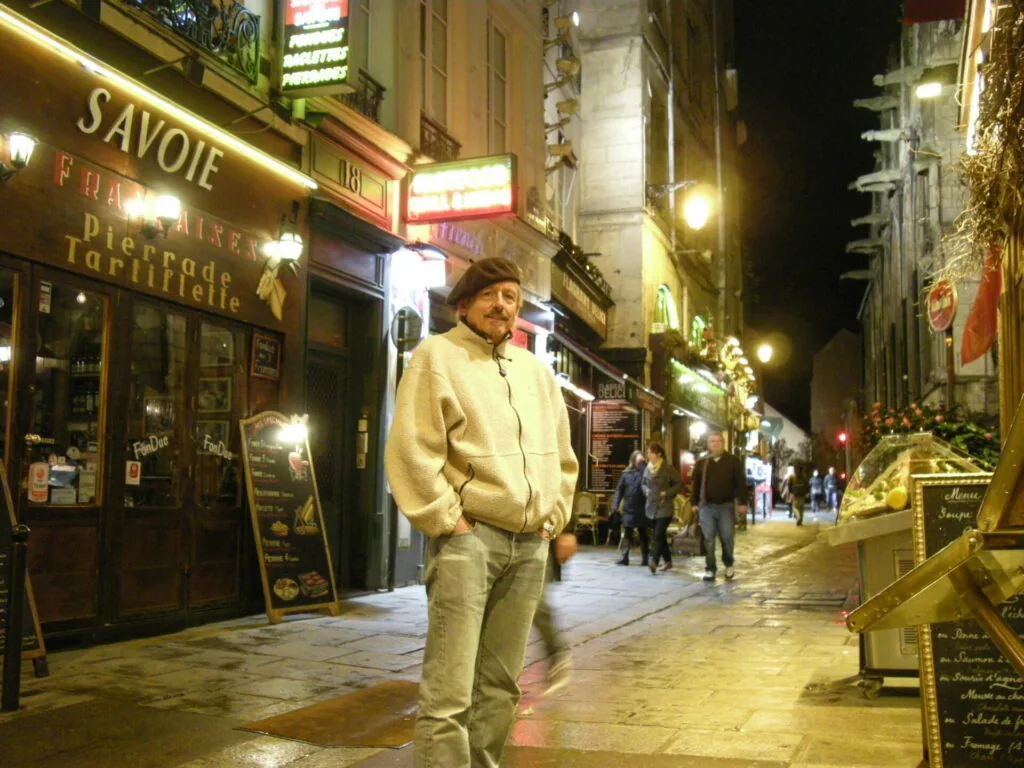 Man in beige jacket standing on illuminated European street at night with restaurant storefronts and pedestrians