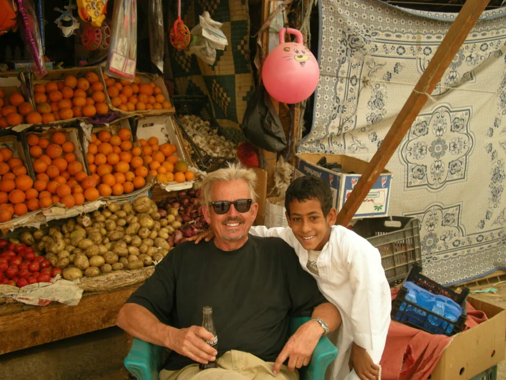 Two men posing in a market stall with baskets of fresh oranges and other produce displayed behind them